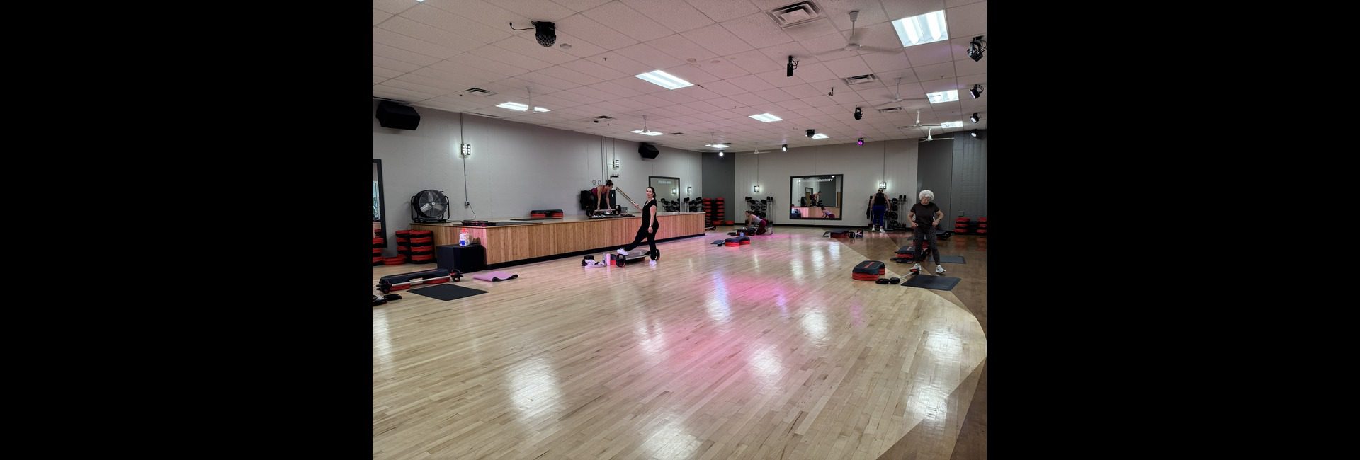 Princeton Club Fitchburg group fitness studio with hardwood floor, instructor platform, wall mirrors, stacked step platforms and mats, with soft pink accent lighting — renovated 2025